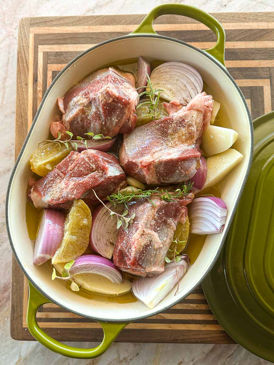 Boneless lamb shanks arranged with potatoes, red onion, garlic and herbs before slow cooking.